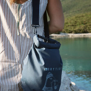 Home 18 Waterproof dry bag with Drytide logo hanging from a person's shoulder near a river.